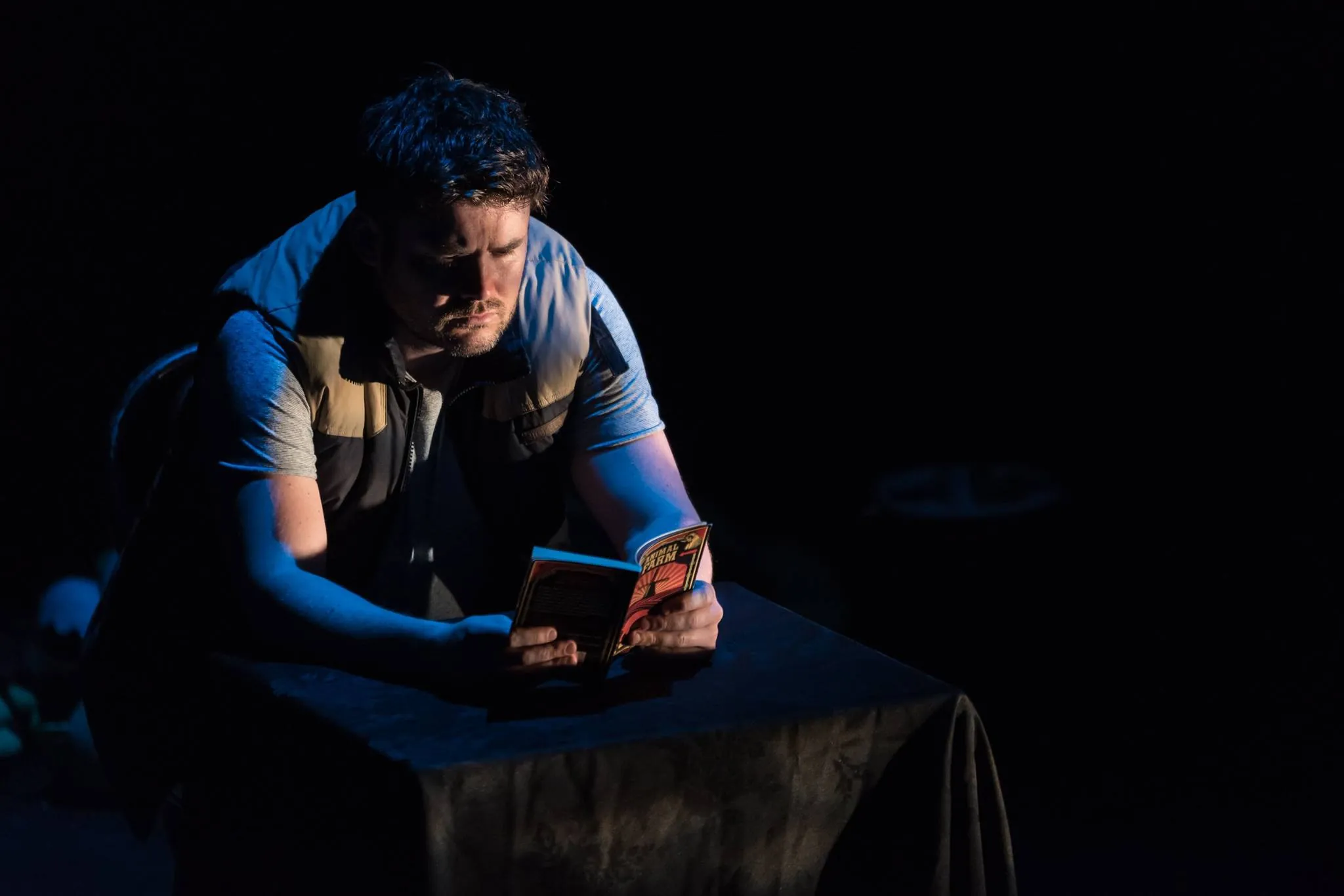 Actor seated at a small table reading a paperback, lit by a single spotlight in darkness