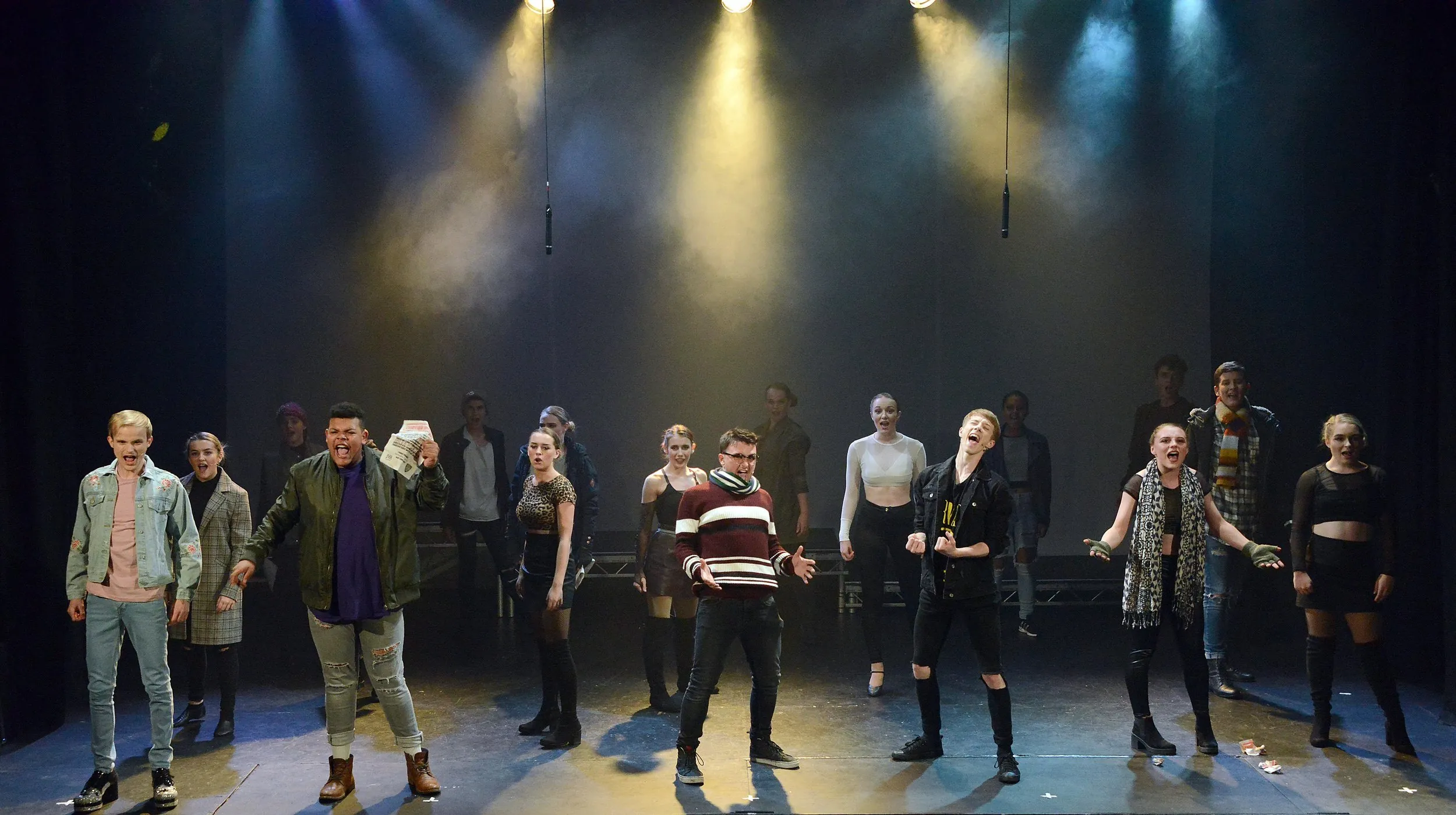 Large youth cast in modern dress singing downstage under hazy yellow and blue backlight