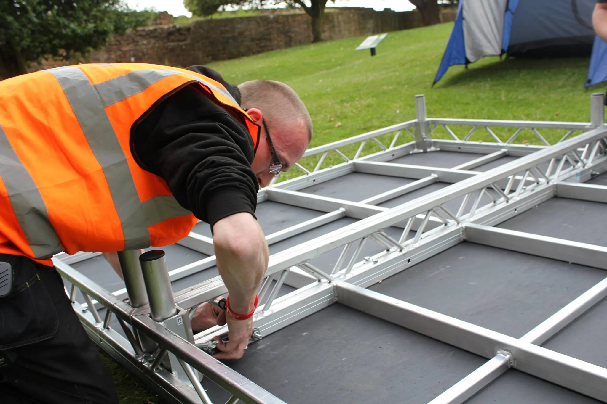 Crew member in orange hi-vis assembling an outdoor stage deck from aluminium truss on a lawn