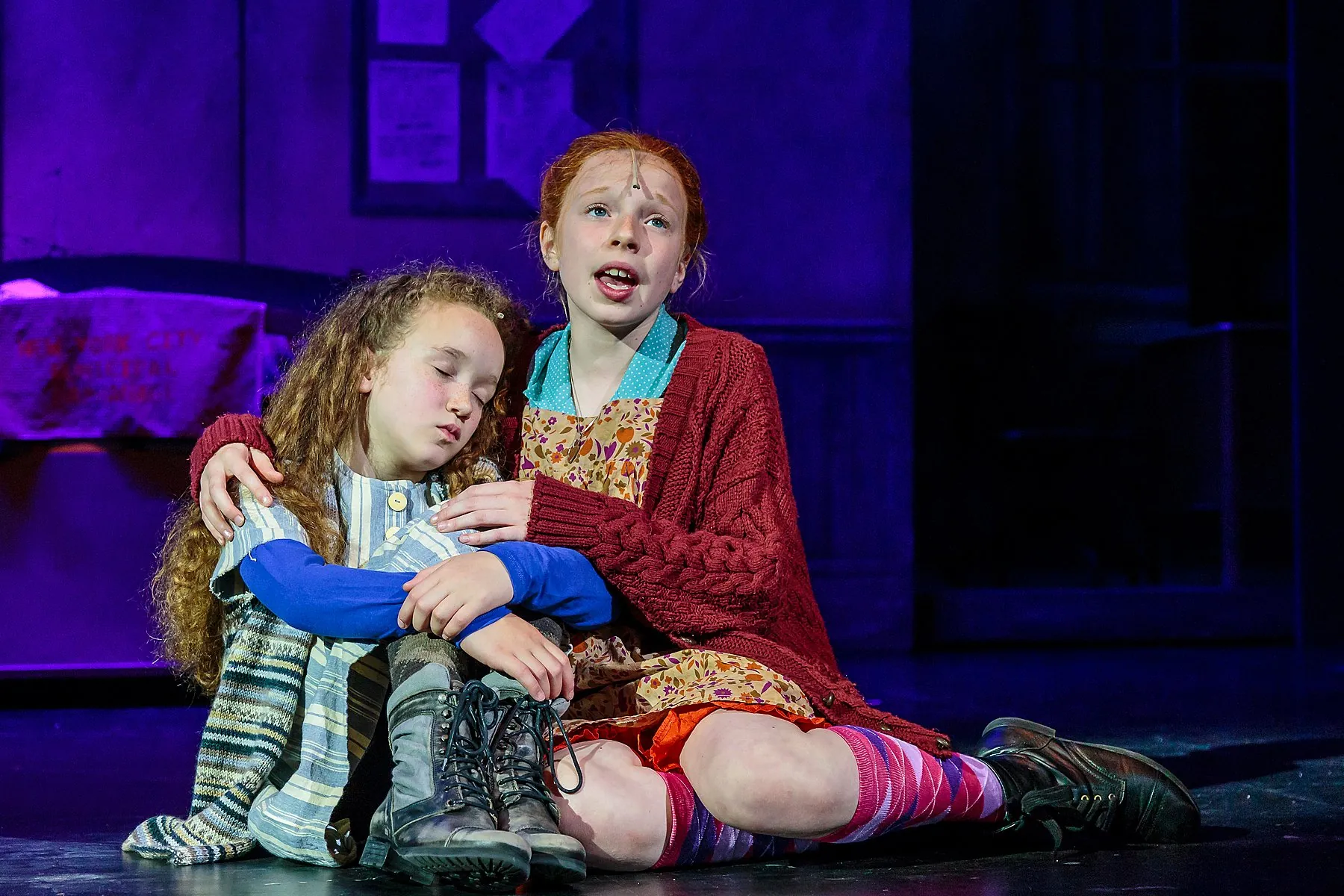 Two young actors performing in Annie, one comforting a sleeping orphan on a purple-lit dormitory set