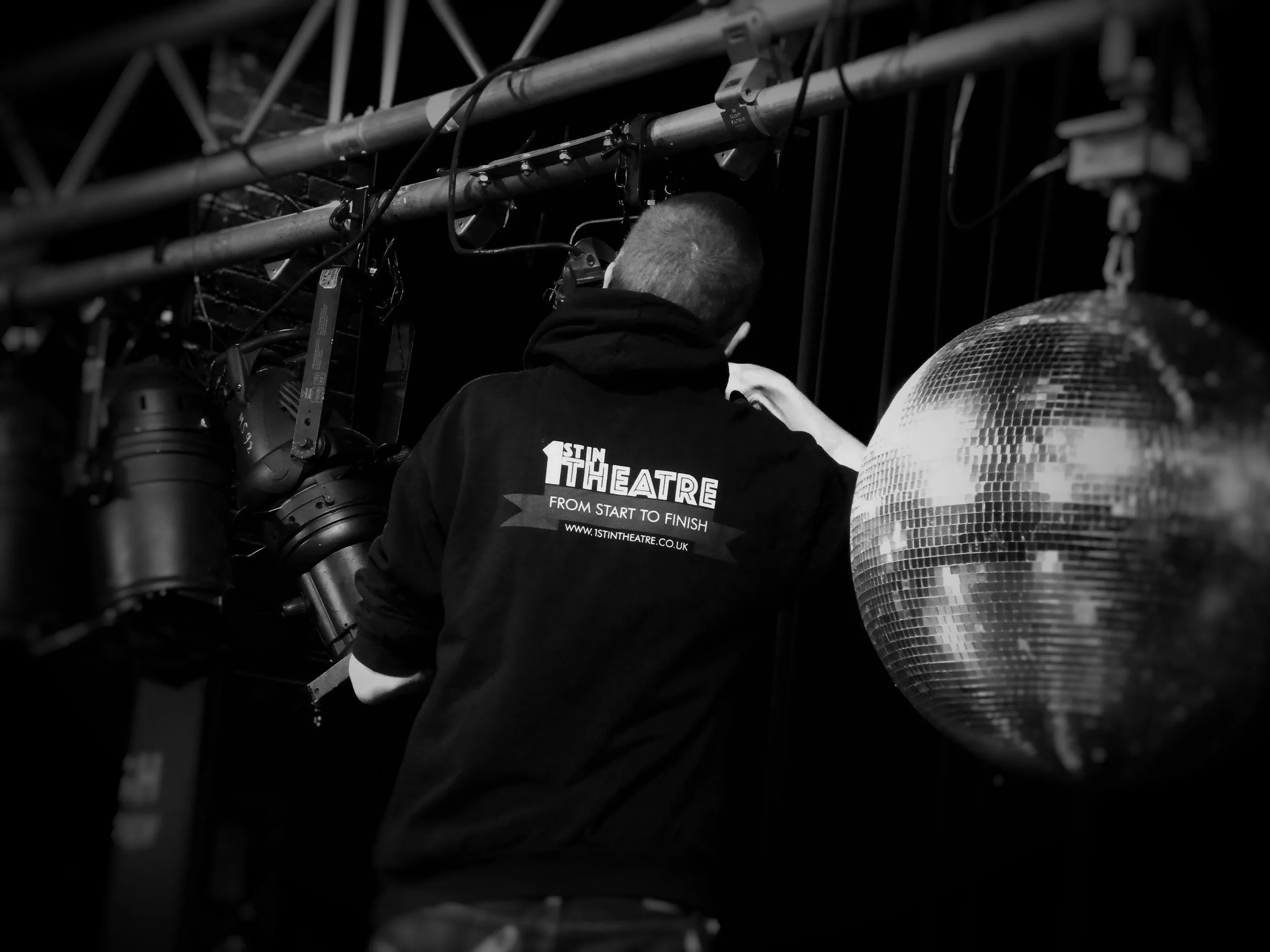 1stintheatre technician adjusting stage lanterns on a lighting bar beside a mirror ball, in black and white