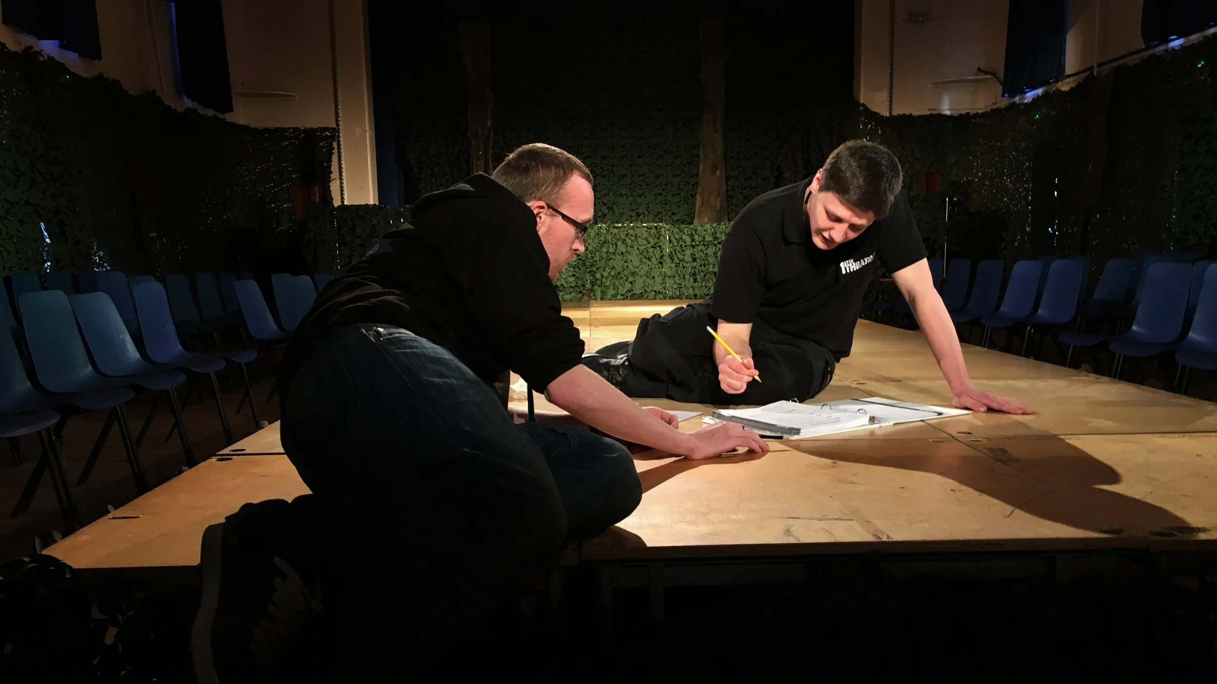 Two crew members kneeling on a bare stage in a hall, marking up plans during a production fit-up