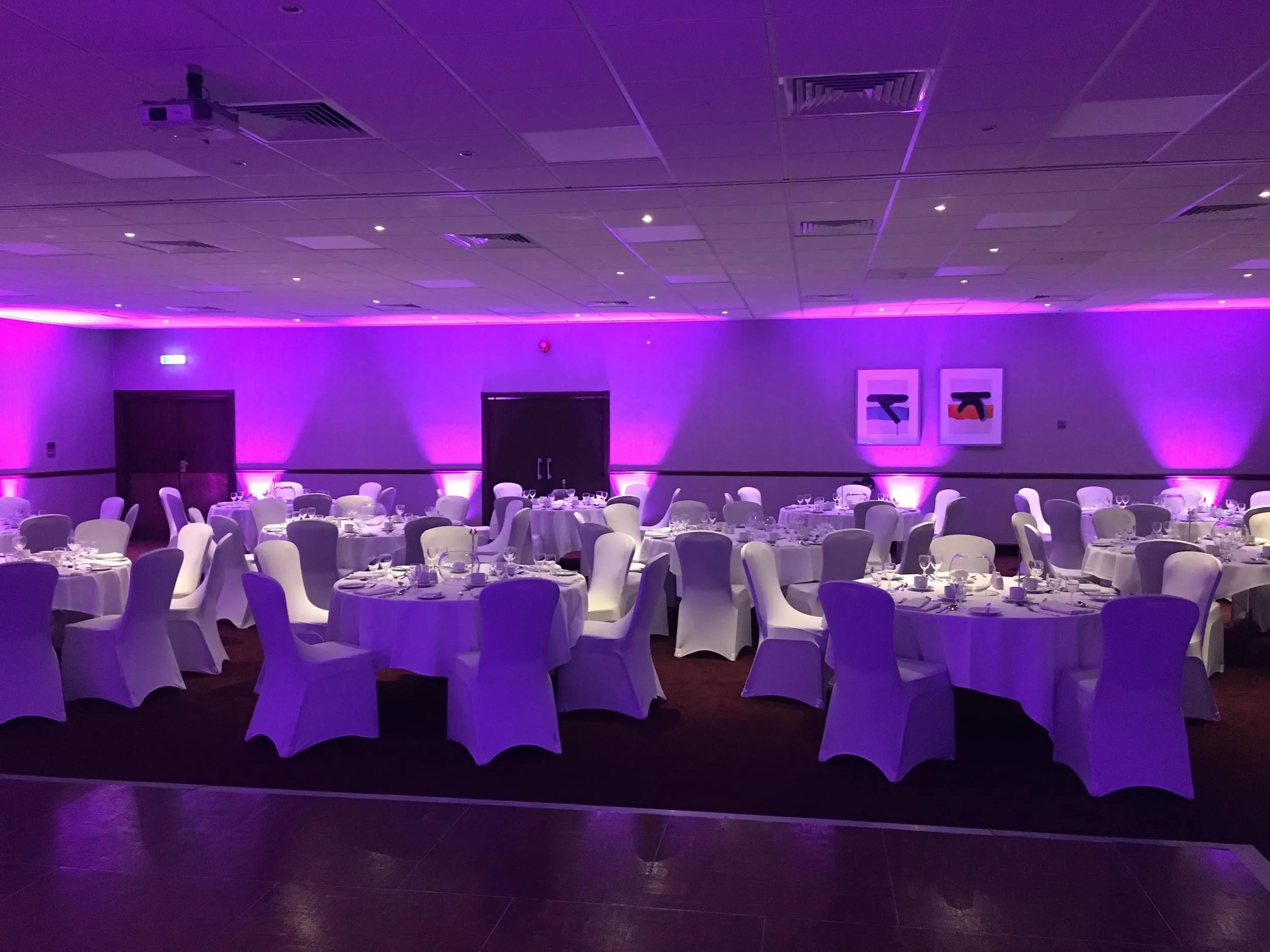 Banqueting room set with round tables and white chair covers, walls washed in purple uplighting
