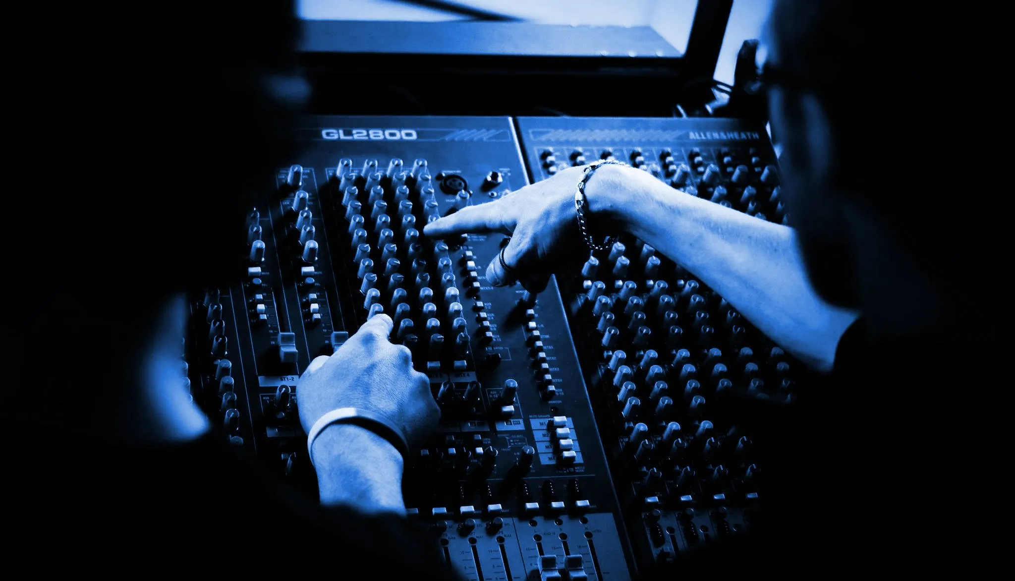 Trainer pointing out channel controls on an Allen & Heath mixing desk to a student, in blue tones