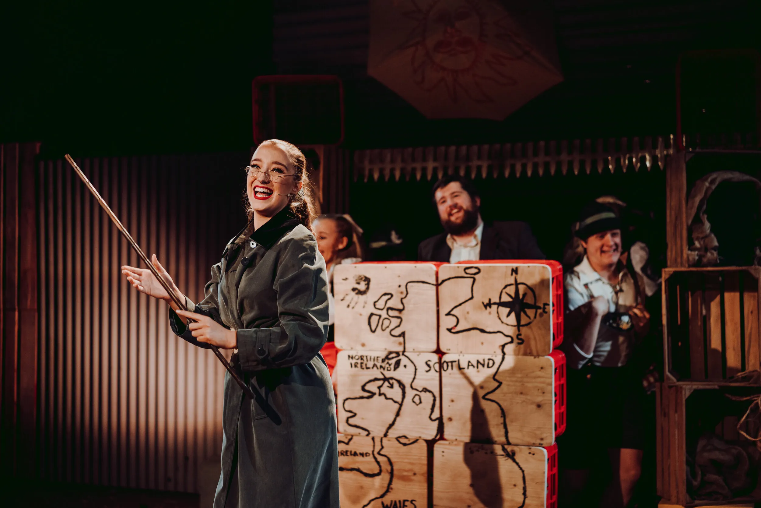 Actress with a pointer stick laughing on stage beside crates painted with a map of Britain, cast smiling behind