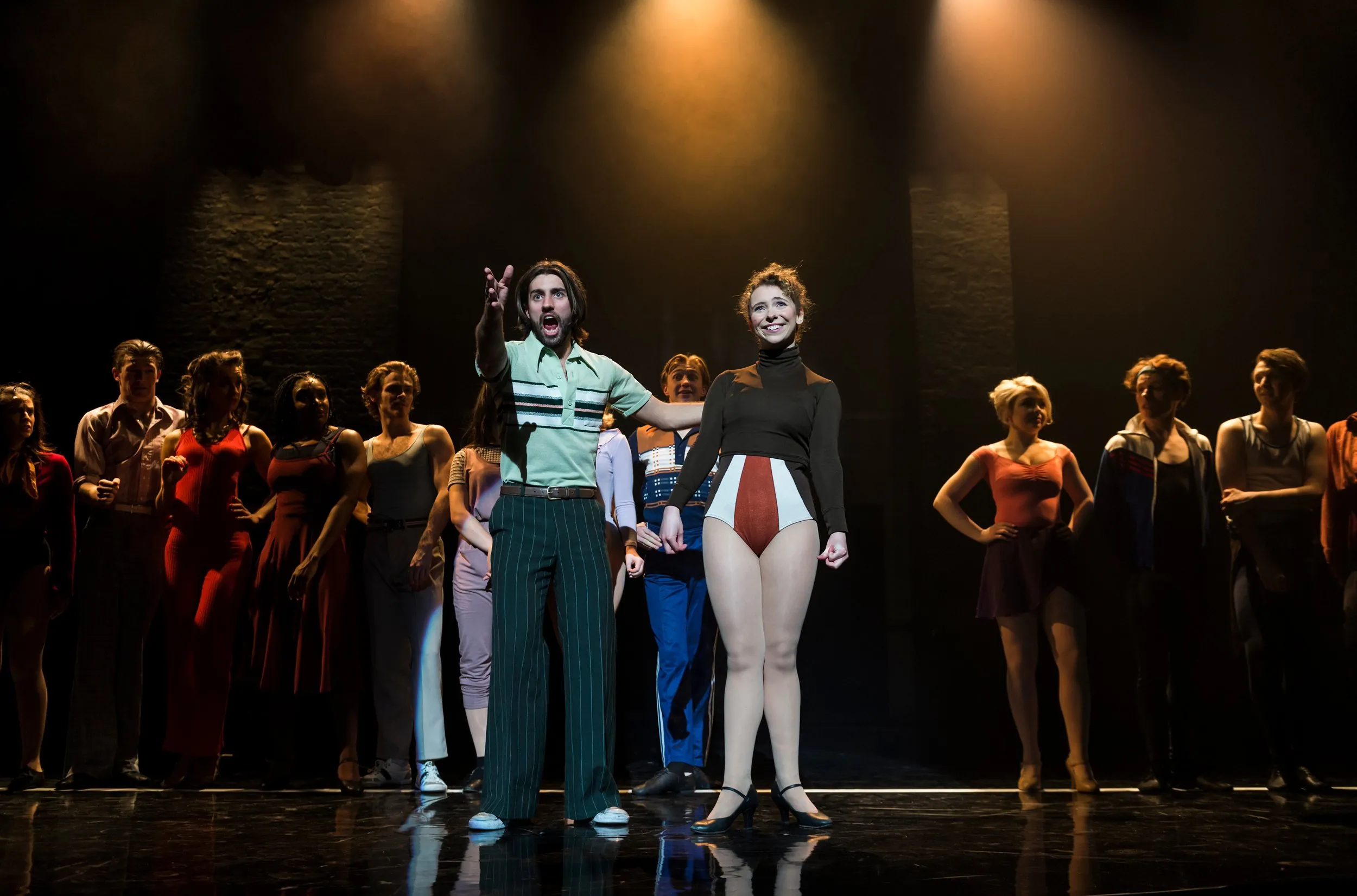 Two lead performers in 1970s costume centre stage with a line of dancers behind under warm spotlights