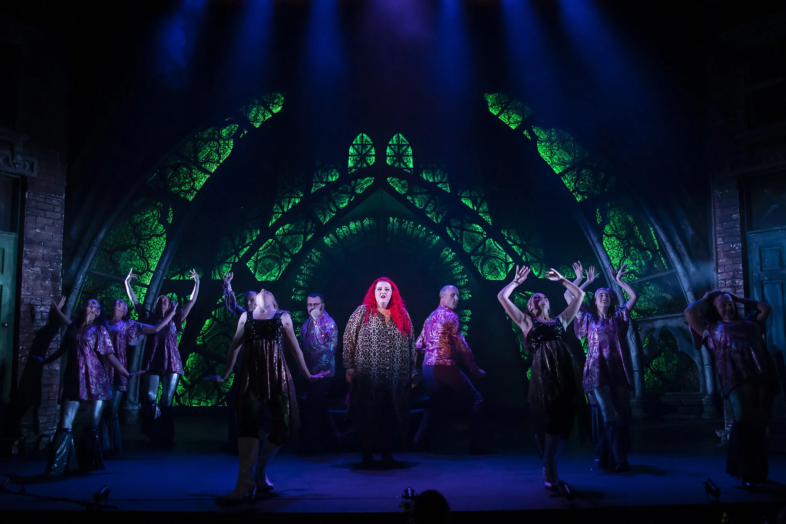 Musical ensemble in shimmering costumes, lead singer centre stage before glowing green gothic arch windows